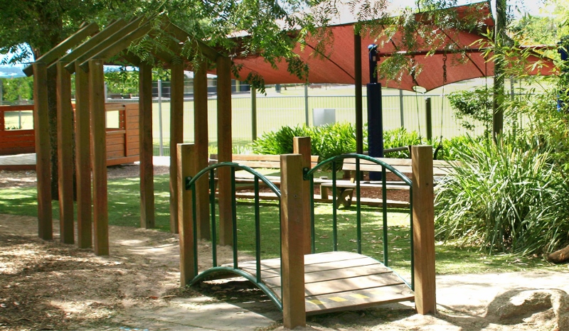Little play bridge in Estrella's outdoor play area, leading onto a path surrounded by wooden archways, beautiful sunny day, with lush green grass, park benches and a big shade cloth nearby.